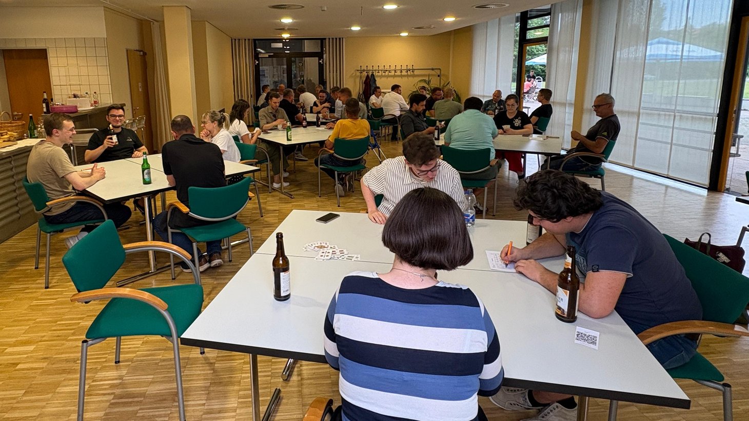 Teilnehmende spielen Skat an mehreren Tischen in einem hellen Veranstaltungsraum. Karten, Getränke und Wertungszettel liegen auf den Tischen, während konzentriert gespielt und Ergebnisse notiert werden.