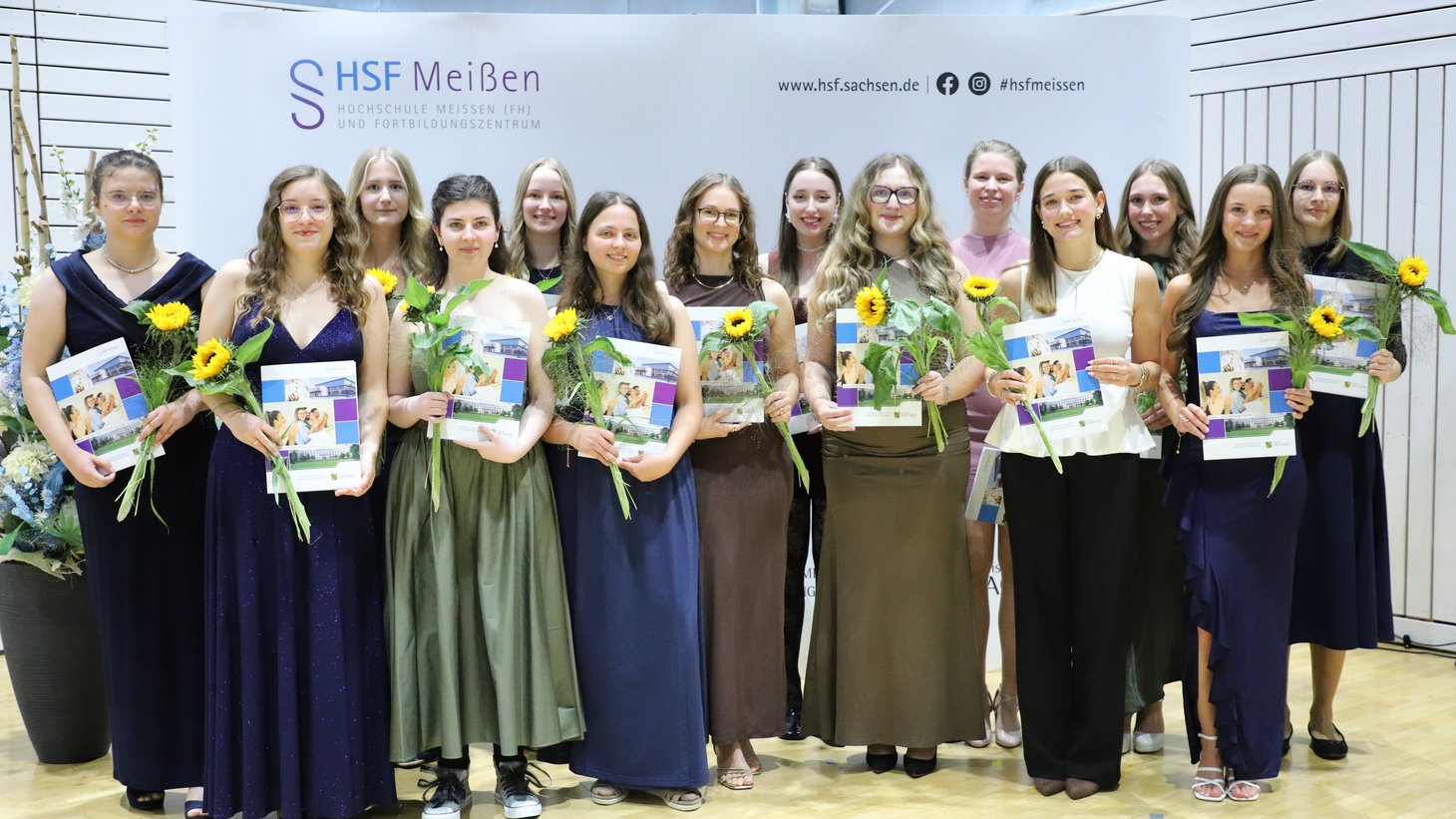 Festlich gekleidete Absolventinnen des Studiums "Sozialverwaltung" stehen lächeln vor der Fotowand in der Mehrzweckhalle der HSF Meißen und halten ihre Zeugnisse und Blumen in den Händen.