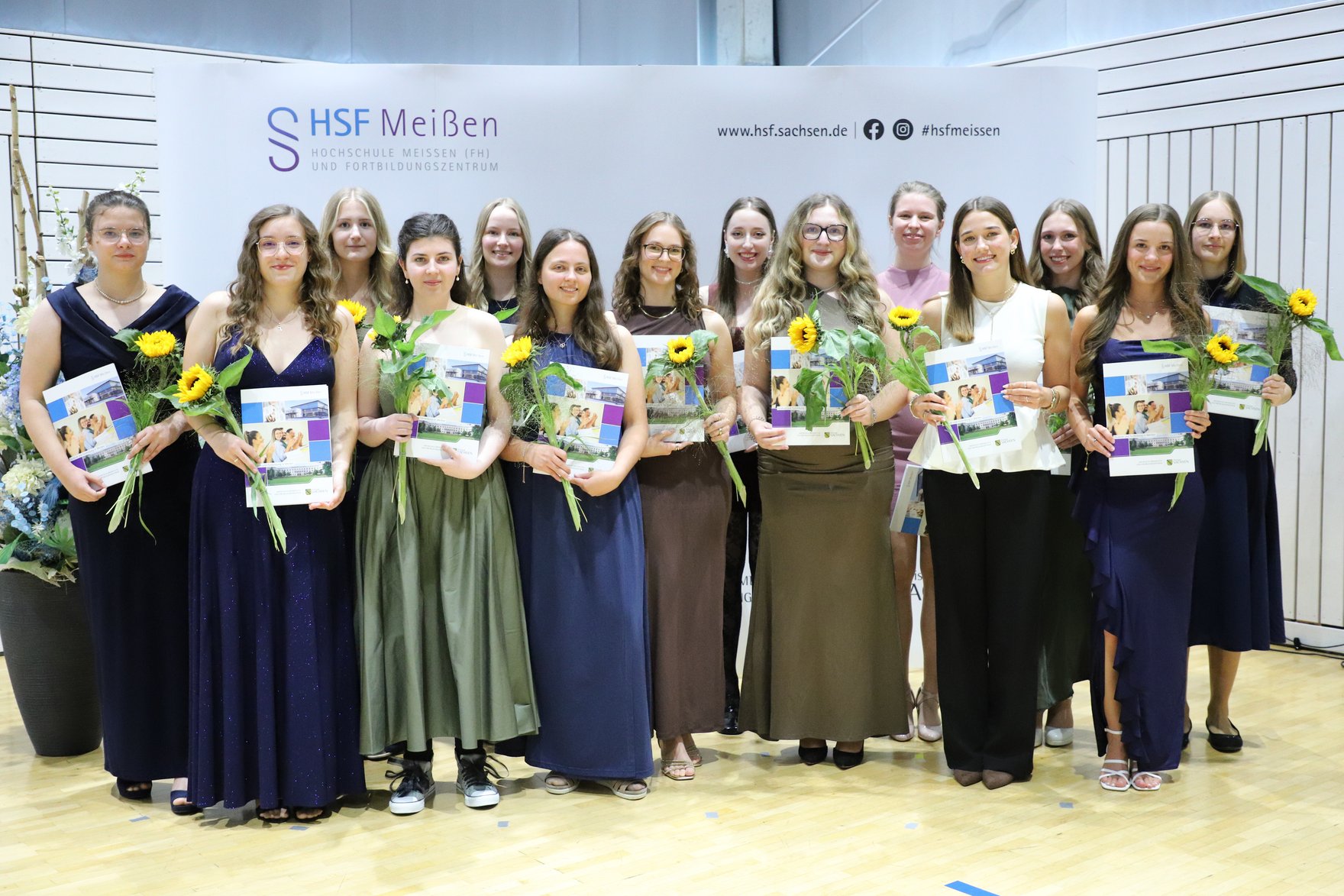 Absolventinnen des Studiums "Sozialverwaltung" Festlich gekleidete Absolventinnen des Studiums "Sozialverwaltung" stehen lächeln vor der Fotowand in der Mehrzweckhalle der HSF Meißen und halten ihre Zeugnisse und Blumen in den Händen.