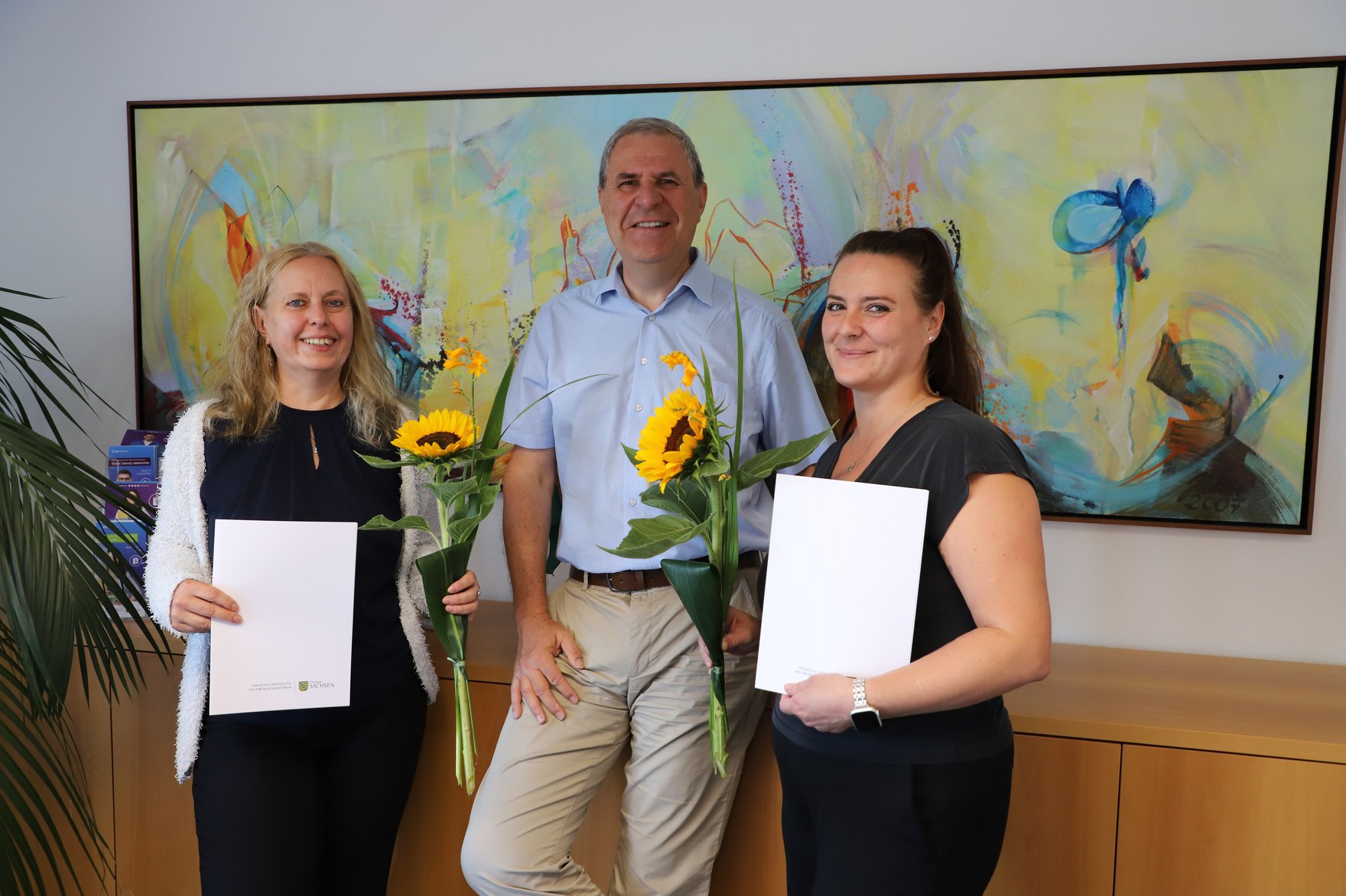 Der Rektor (Mitte) überreicht einer Hochschulmitarbeiterin (rechts) und einer Dozentin (link) die Bestellungsurkunden als Konflikt- und Mobbingberaterinnen. Die beiden stehen links und rechts neben ihm, halten ihre Urkunden sowie jeweils eine Sonnenblume in den Händen und lächeln in die Kamera. Im Hintergrund hängt ein großformatiges, farbenfrohes abstraktes Gemälde.