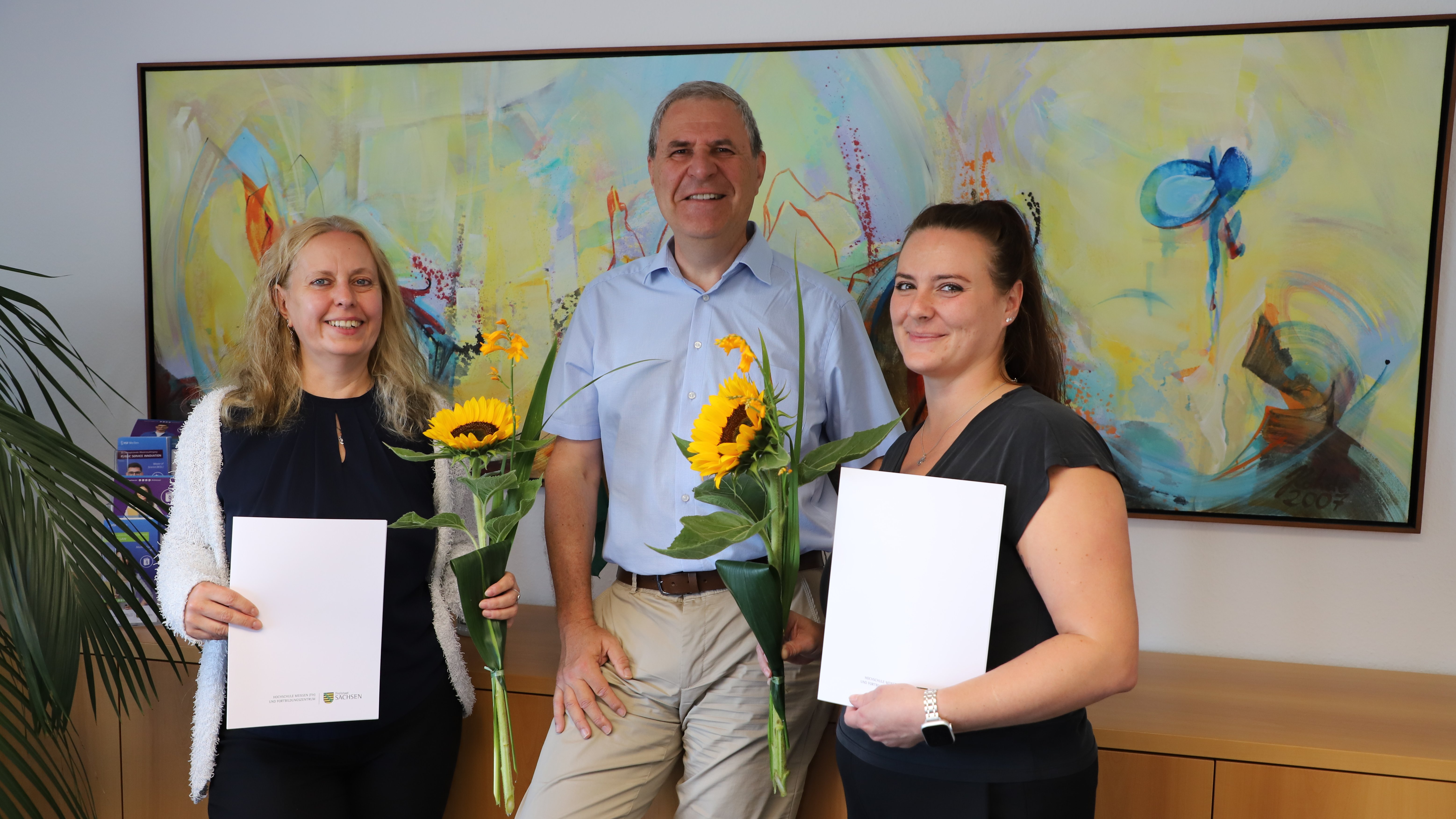 Der Rektor (Mitte) überreicht einer Hochschulmitarbeiterin (rechts) und einer Dozentin (link) die Bestellungsurkunden als Konflikt- und Mobbingberaterinnen. Die beiden stehen links und rechts neben ihm, halten ihre Urkunden sowie jeweils eine Sonnenblume in den Händen und lächeln in die Kamera. Im Hintergrund hängt ein großformatiges, farbenfrohes abstraktes Gemälde.