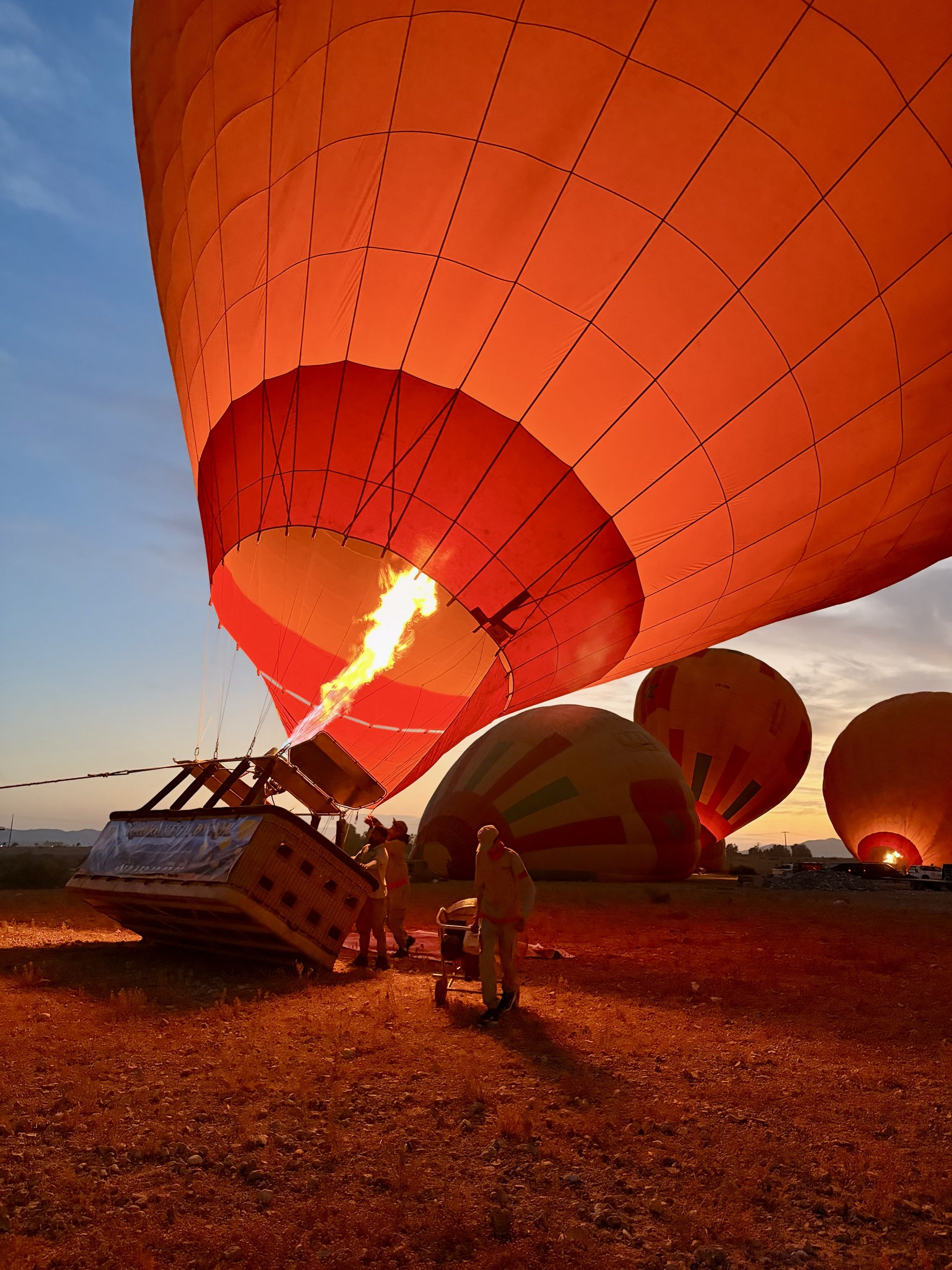 Ein Heißluftballon wird in den frühen Morgenstunden mit heißer Luft befüllt, während die Crew den Korb vorbereitet. Im Hintergrund sind weitere Ballons zu sehen, die für den Start bereitstehen.