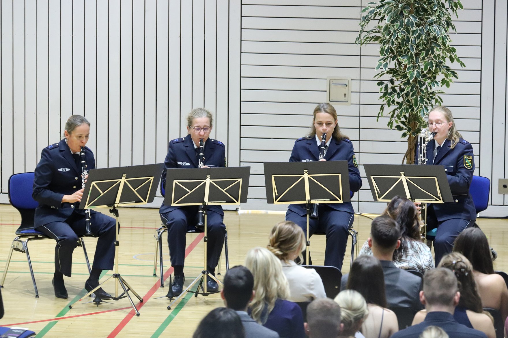 Klarinettenquartett des Sächsischen Polizeiorchesters Auf vier Stühlen sitzt vor den Gästen der Zeugnisübergabe das Klarinettenquartett des Sächsischen Polizeiorchesters. Die vier Frauen spielen derzeit ein musikalisches Stück. Alle tragen die dunkelblaue Polizeiuniform.
