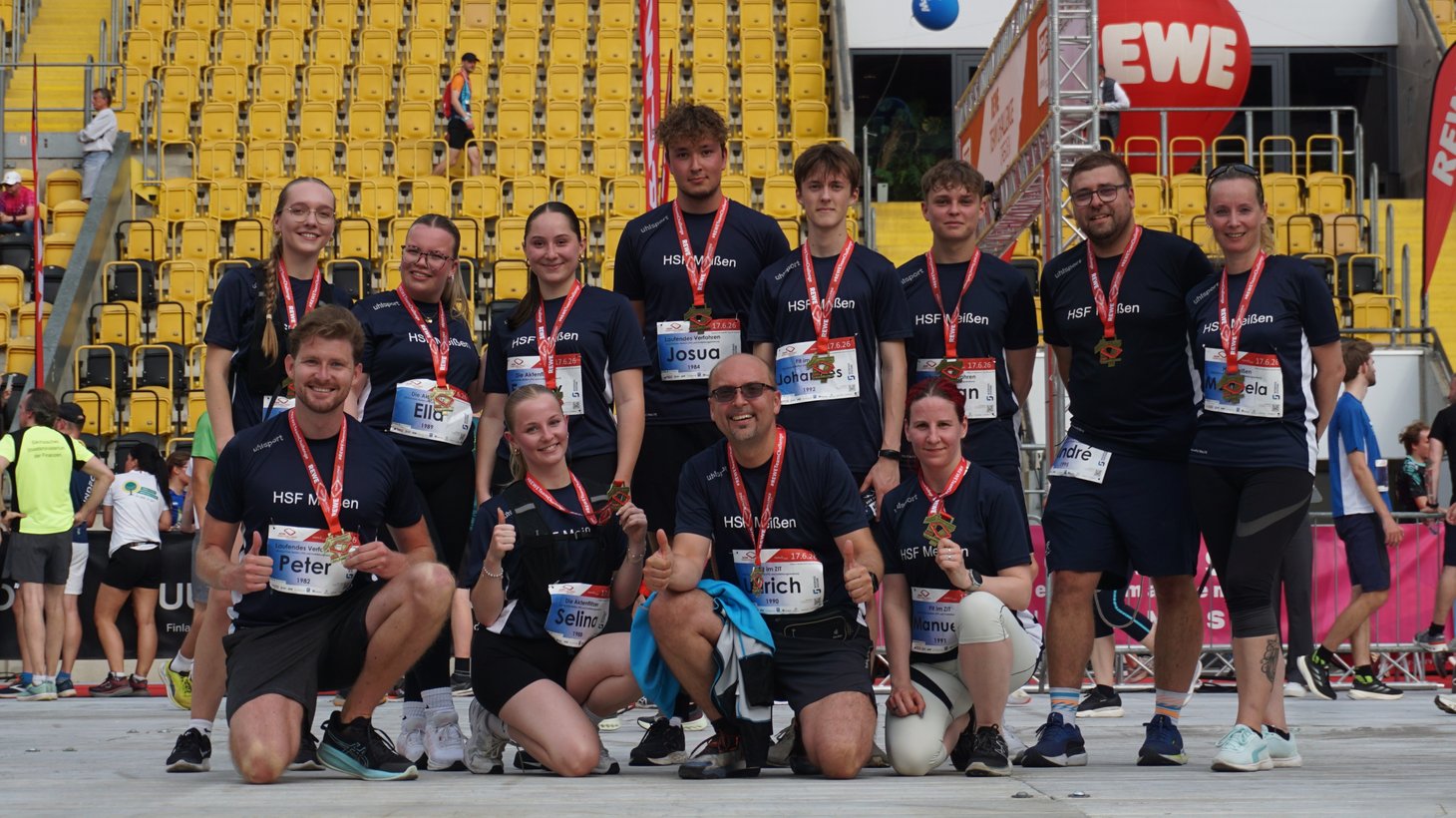 Das Team „HSF Meißen“ posiert nach der REWE Team Challenge Dresden gemeinsam für ein Gruppenfoto im Stadion. Alle Teilnehmenden tragen ihre Finisher-Medaillen und lächeln in die Kamera, einige zeigen den Daumen nach oben. Im Hintergrund sind die gelben Tribünen des Stadions sowie der Zielbereich der Veranstaltung zu sehen.